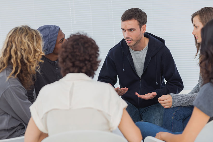 Group of six young men and women seated and having a discussion in a group therapy setting.